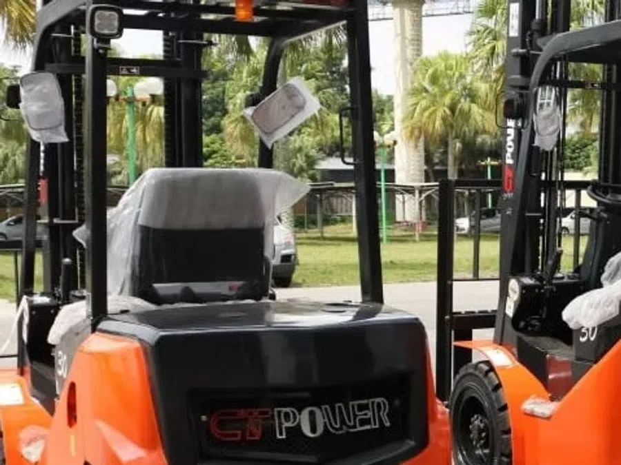 Row of orange forklifts parked outdoors with palm trees and greenery in the background under a clear sky