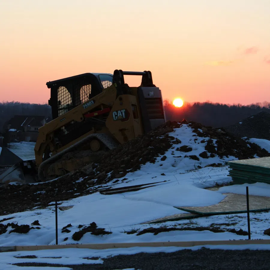 CAT skid steer loader working on a snowy construction site during sunset with houses in the background