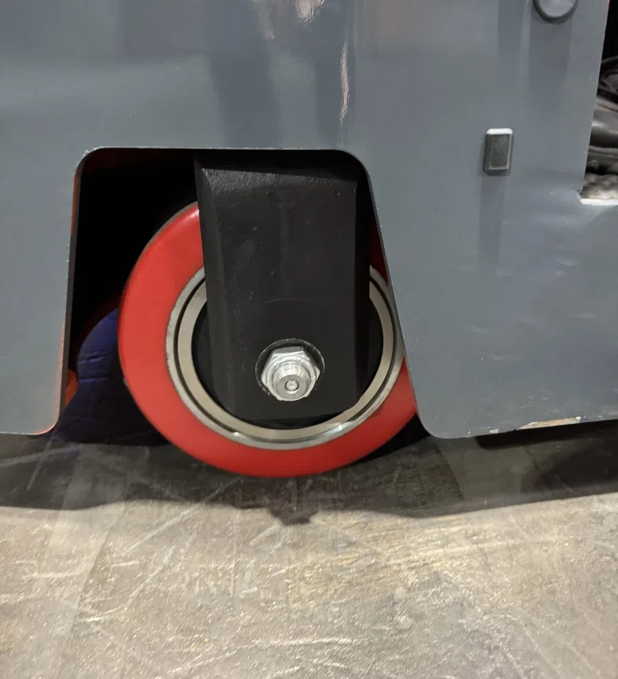 Close-up of a red caster wheel with metal bolt attached to a gray industrial machine or equipment.