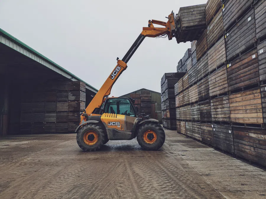 Yellow JCB forklift stacking wooden crates in an industrial warehouse yard on a cloudy day