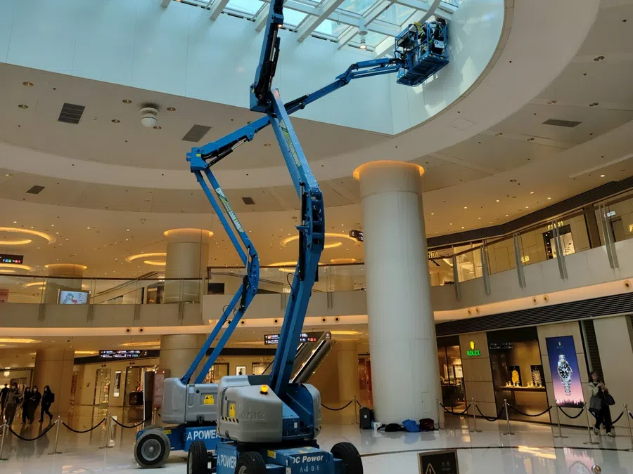 Blue articulated boom lift inside a shopping mall extending to clean or maintain the glass ceiling skylight.