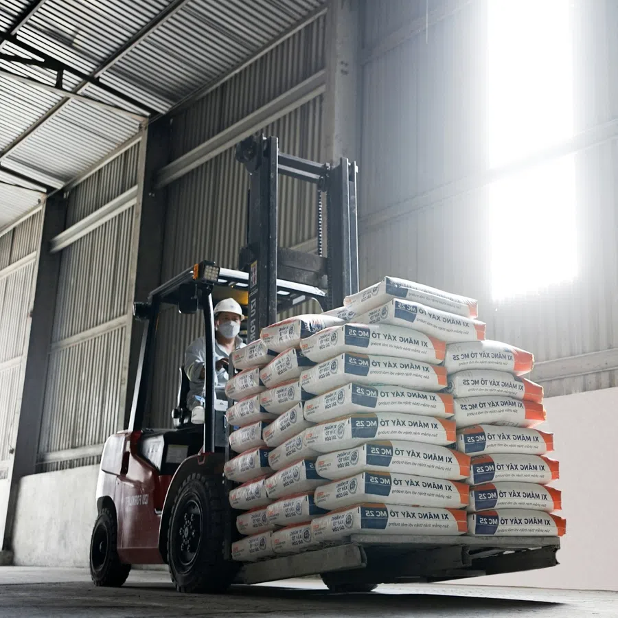 Forklift operator wearing safety helmet and mask loading stacked cement bags inside warehouse with metal walls.
