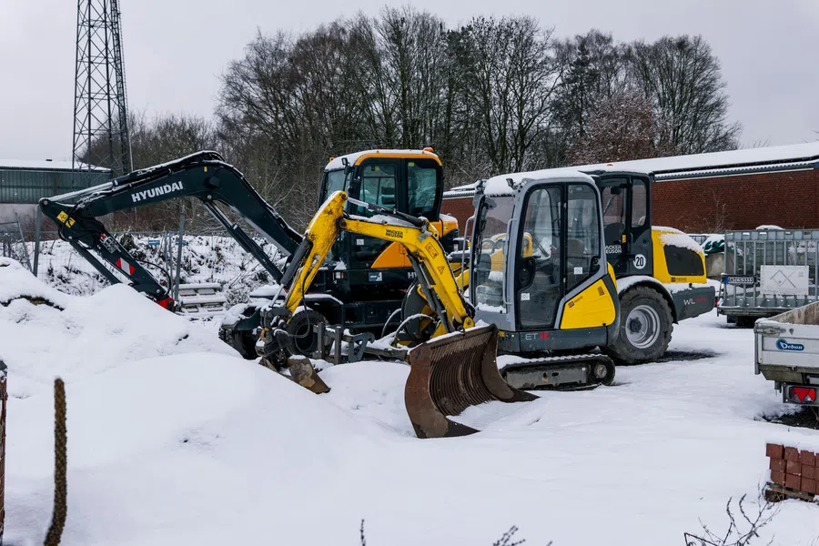 Yellow and black excavators parked on snow-covered ground near construction materials and bare trees in winter.