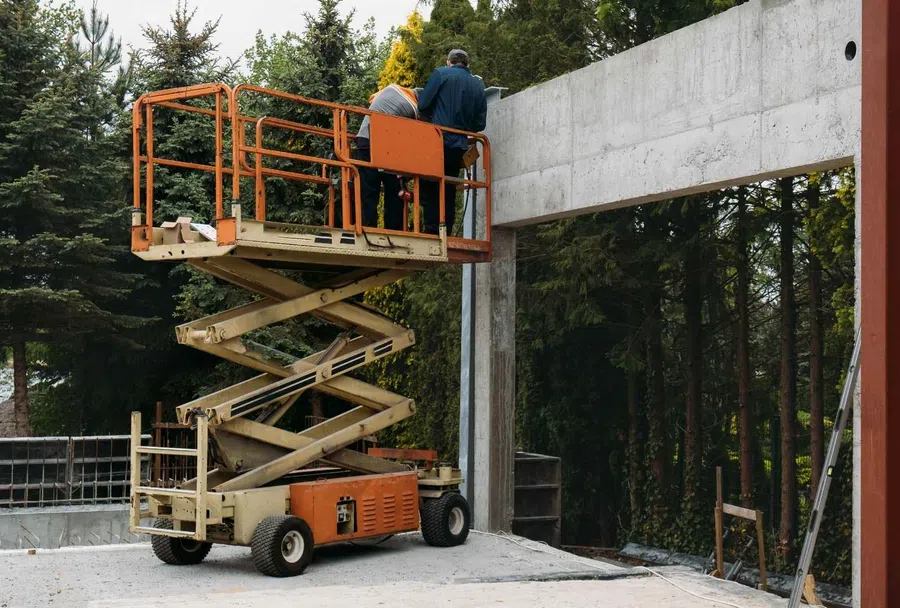 Two workers on an orange scissor lift inspecting a concrete structure at an outdoor construction site surrounded by trees