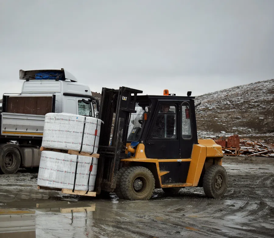 Yellow forklift lifting wrapped pallets in an outdoor muddy industrial area with a white truck in the background.