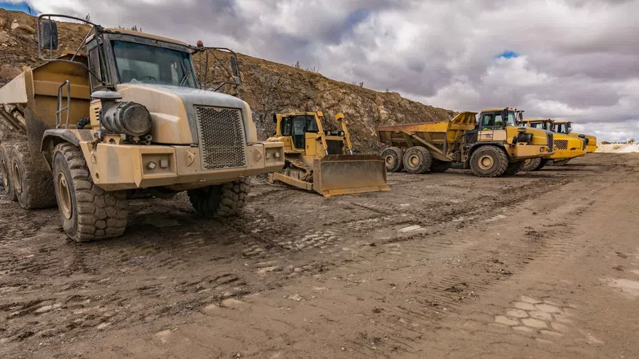 Heavy construction machinery including dump trucks and bulldozer parked on a dirt road at a mining site under cloudy sky