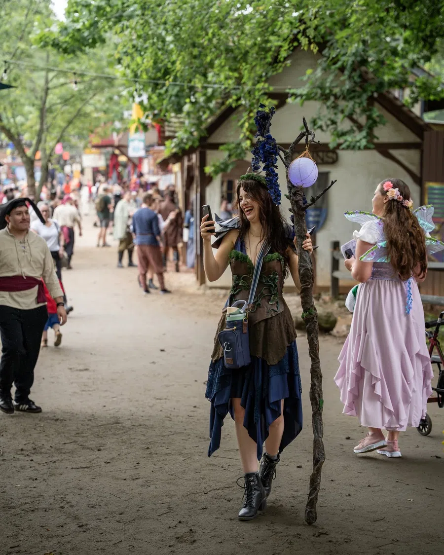 A costumed creator laughing as she films herself on her phone while walking through the Festival village, carrying a fantasy staff.