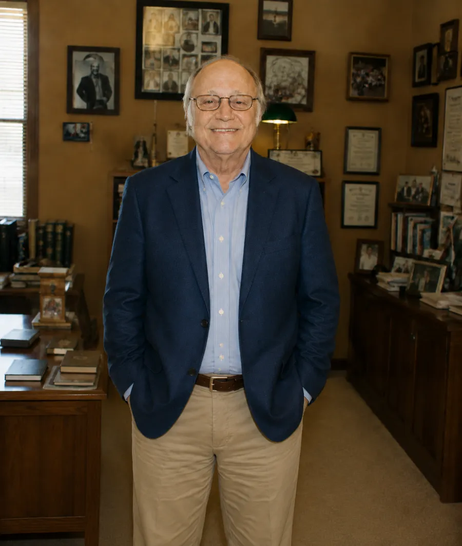 Smiling elderly man in blue blazer and khaki pants standing in a warmly lit, book-filled office with framed pictures on walls.