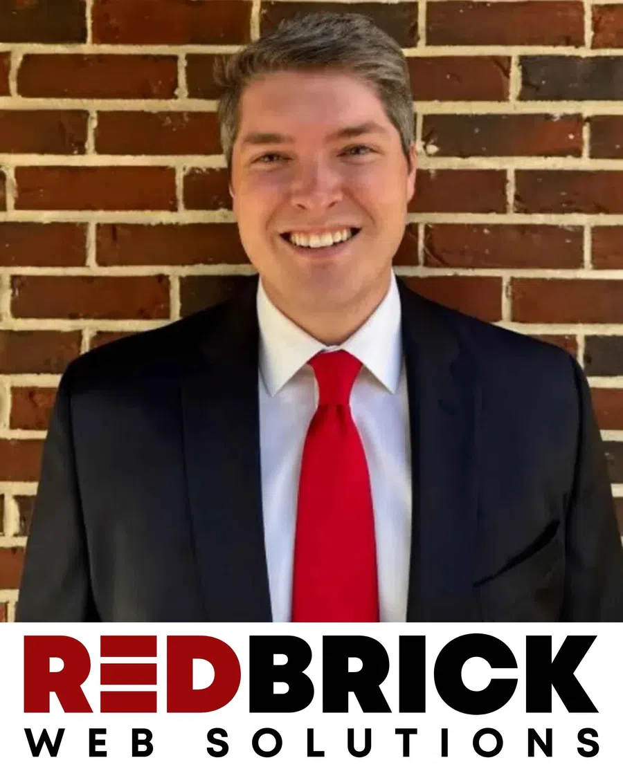 Smiling man in suit and red tie in front of brick wall with RedBrick Web Solutions logo below.