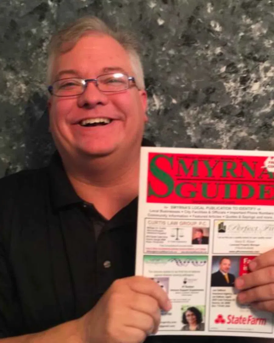 Smiling man with glasses holding a colorful Smyrna Guide publication featuring local ads and guides.