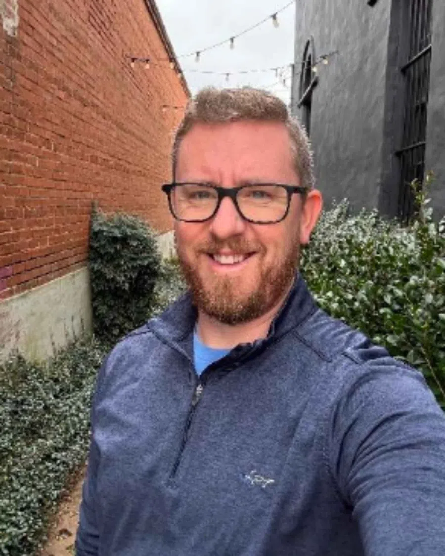 Smiling man with glasses and beard wearing a blue quarter-zip sweater standing outdoors by brick and black walls.