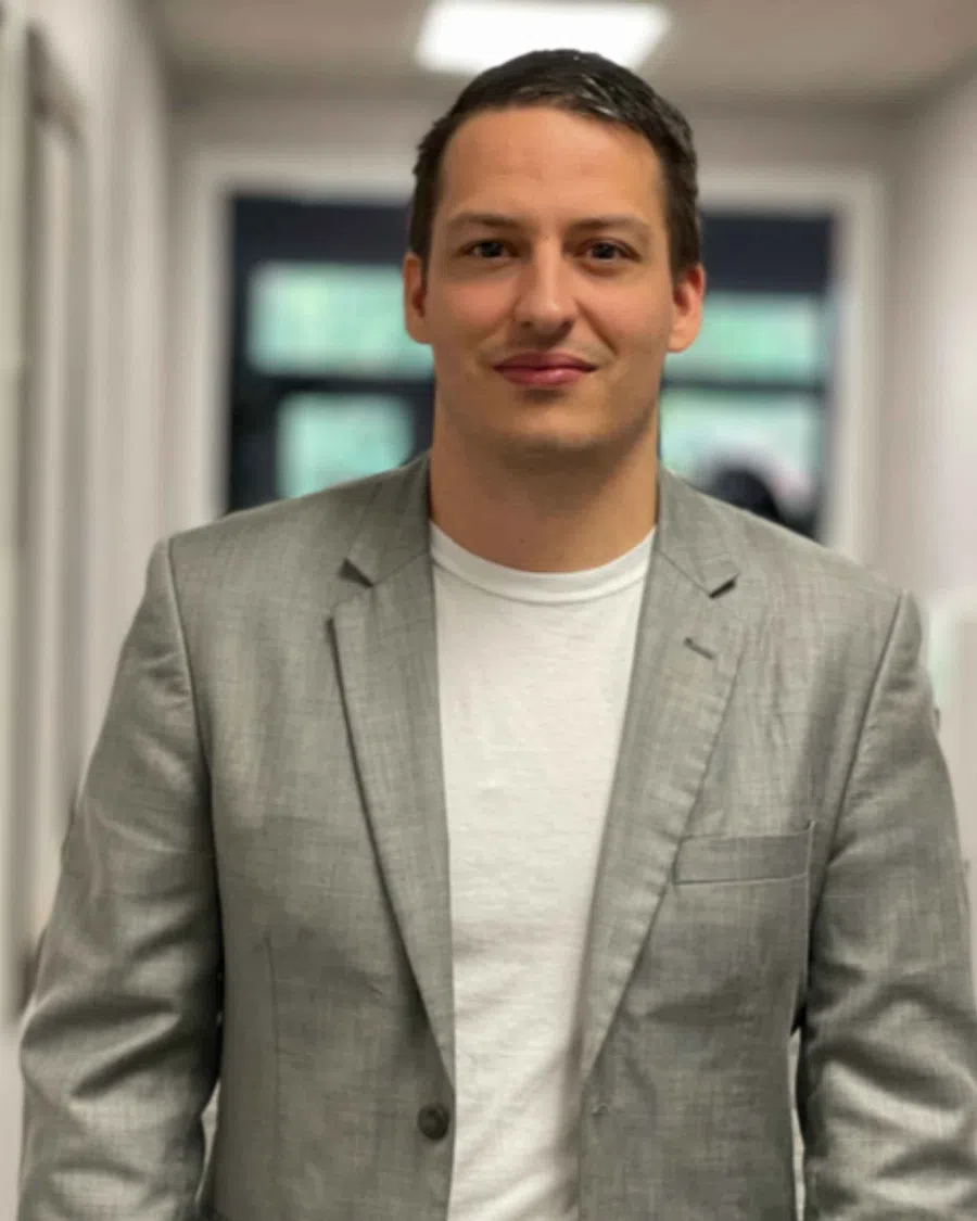 Man in a gray blazer and white t-shirt standing in a hallway with blurred background.