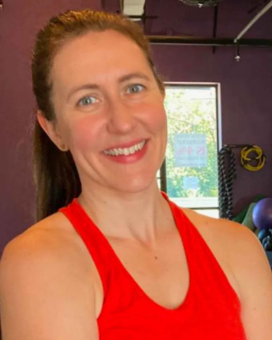 Smiling woman in red tank top standing in a gym with fitness equipment and window in the background