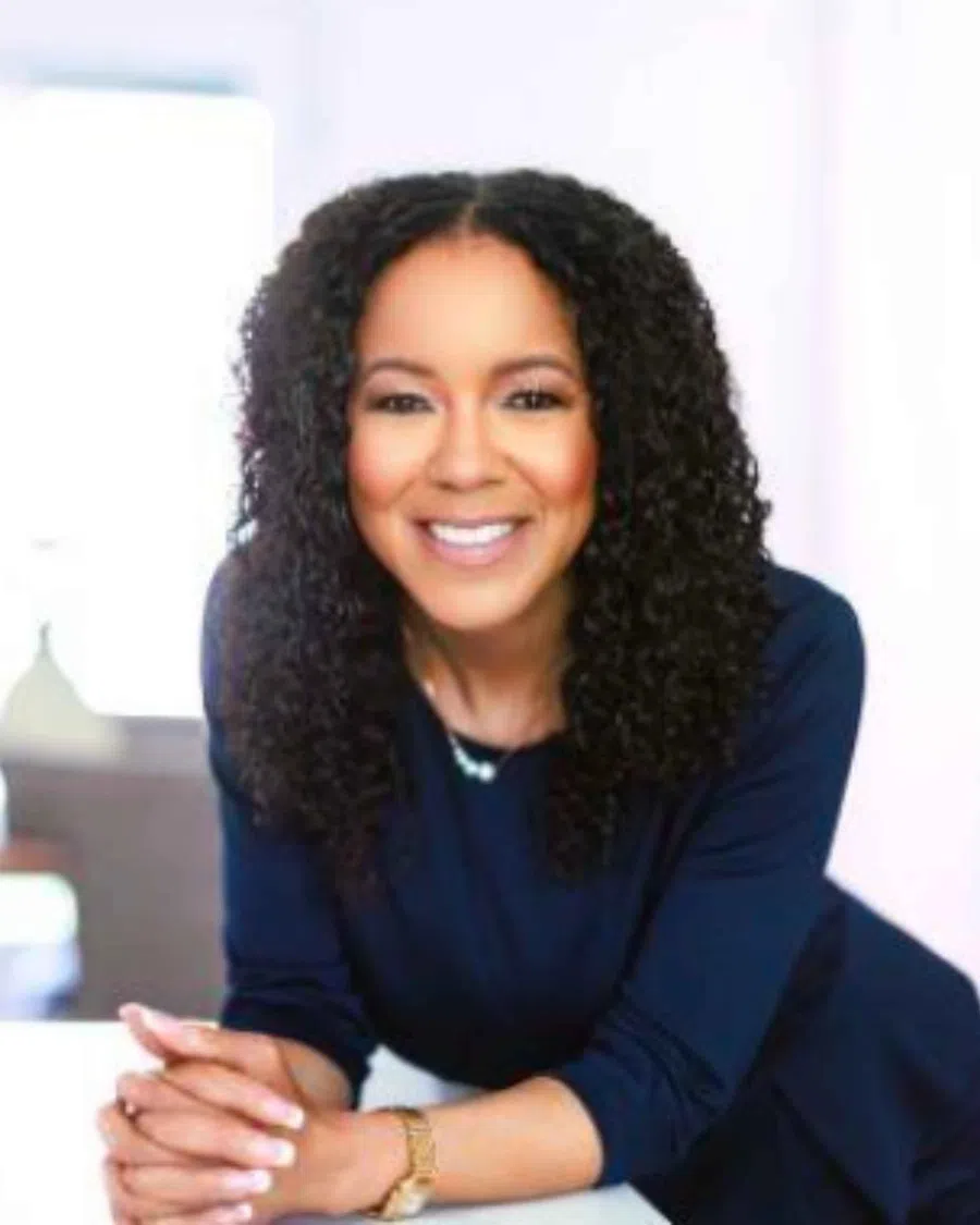 Smiling woman with curly hair in navy top leaning forward with hands clasped in bright indoor setting