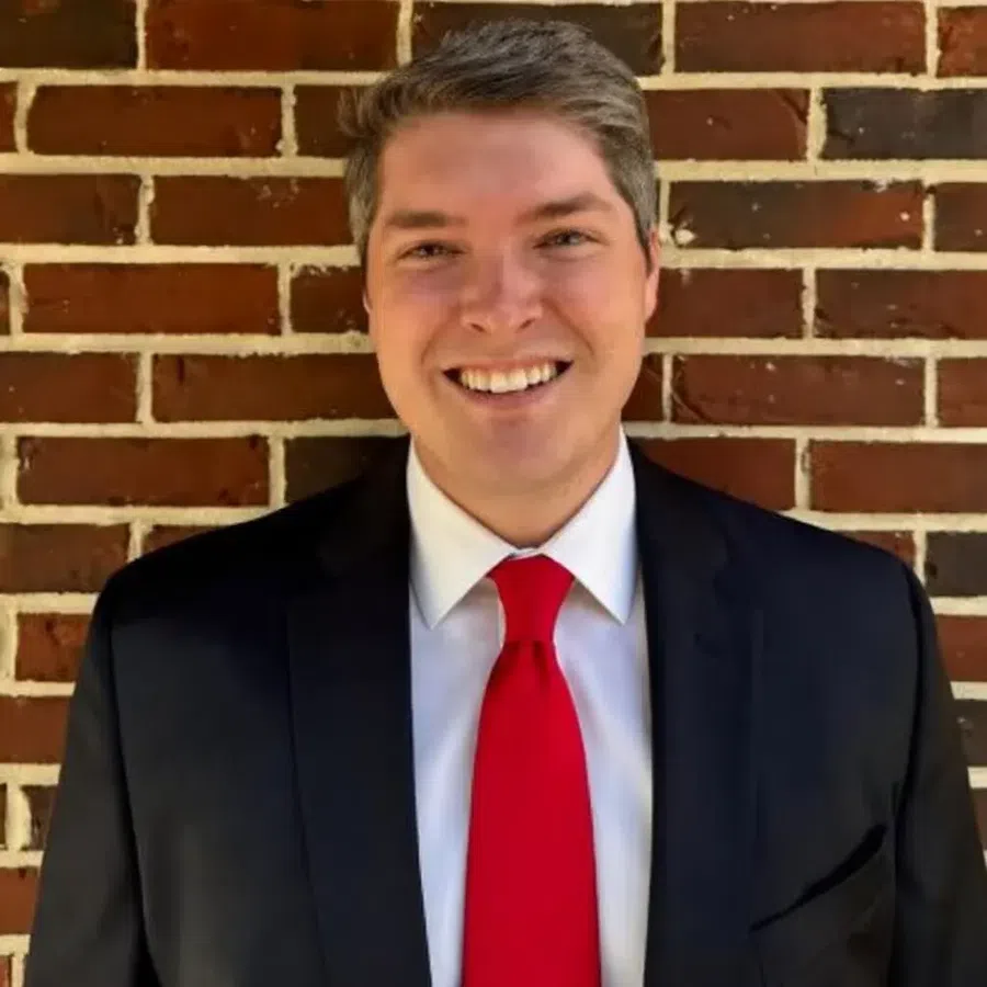 Smiling man wearing a black suit, white shirt, and red tie standing against a brick wall background