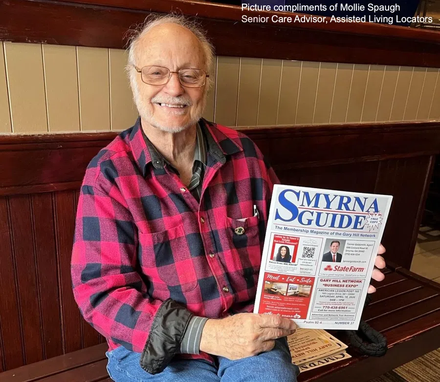 Elderly man in red plaid shirt smiling and holding a Smyrna Guide newsletter while seated on a wooden bench.