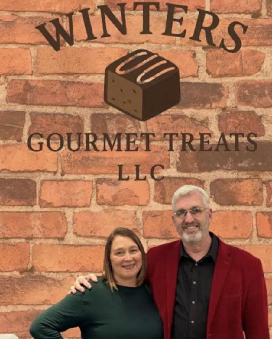Two smiling people stand in front of a brick wall with Winters Gourmet Treats LLC logo featuring a chocolate piece.