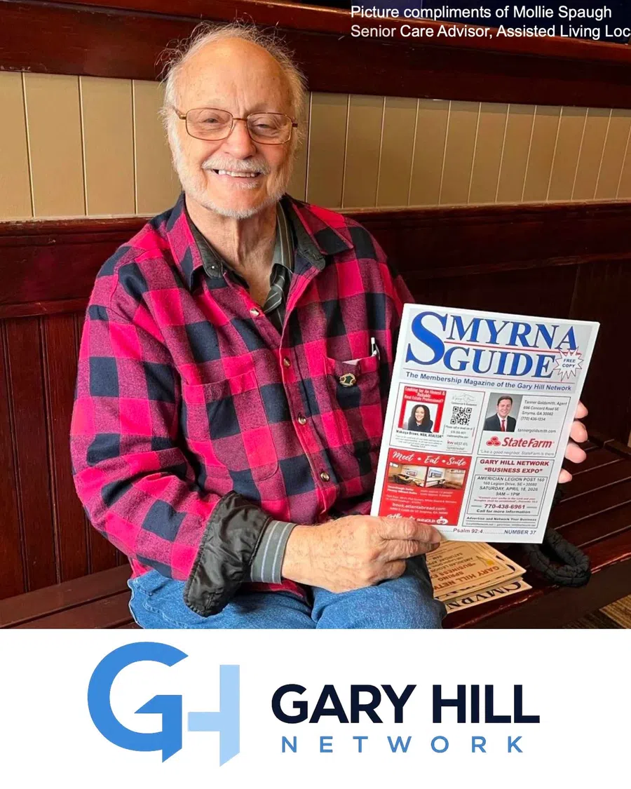 Smiling elderly man in red plaid shirt holding a copy of the Smyrna Guide magazine indoors.