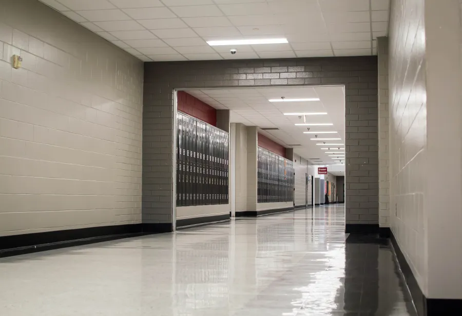 Empty school hallway with shiny floor, beige brick walls, and rows of lockers under fluorescent lights