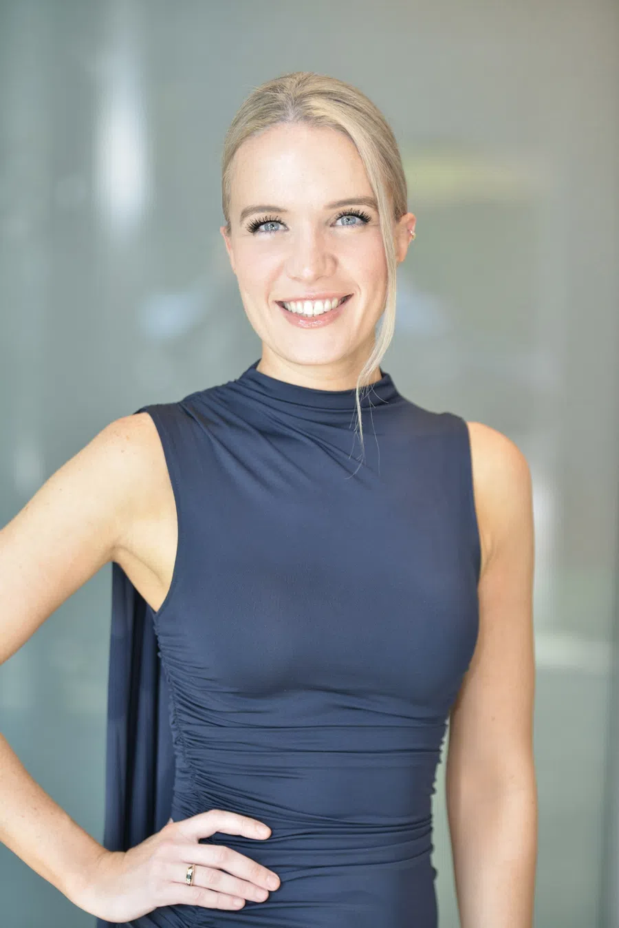 Smiling blonde woman in sleeveless dark blue dress posing with hand on hip against blurred background