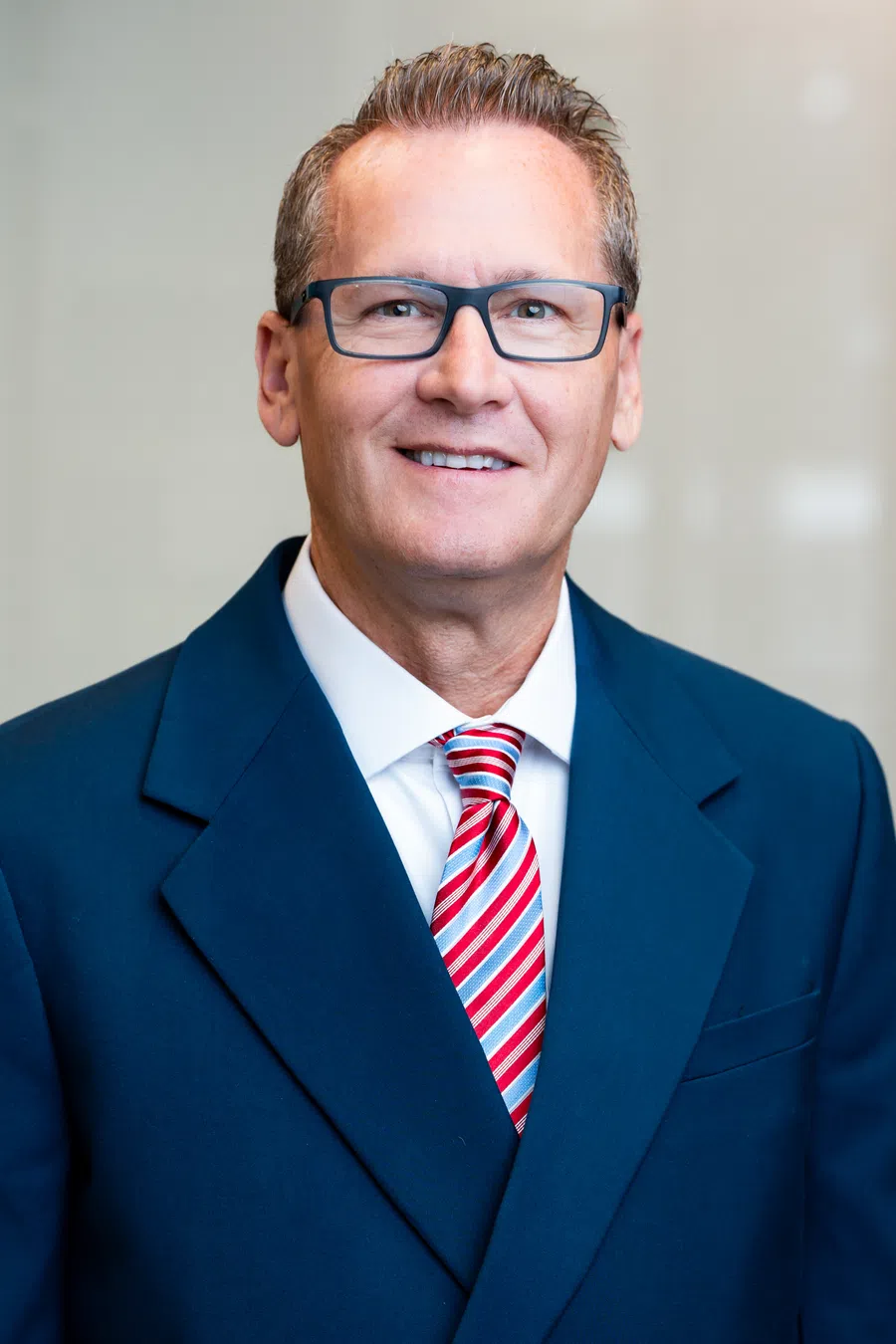 Professional middle-aged man wearing glasses, blue suit, and red striped tie smiling in a corporate setting.