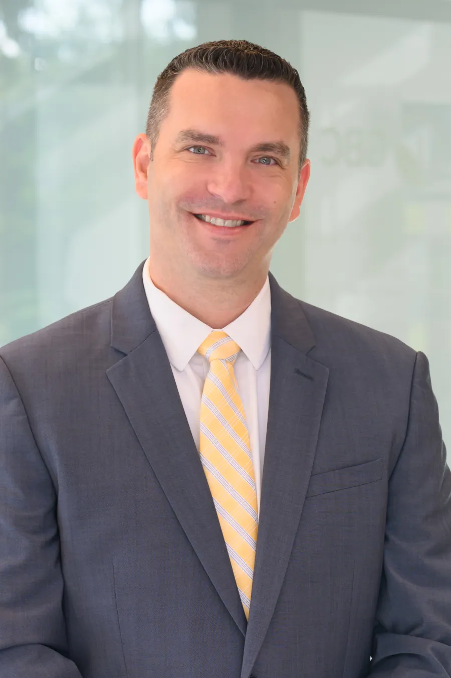 Smiling businessman in gray suit and yellow striped tie standing against a blurred office background.