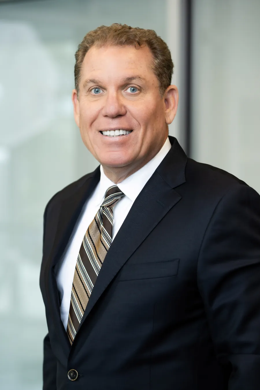 Smiling businessman in black suit and striped tie posing in bright modern office setting.