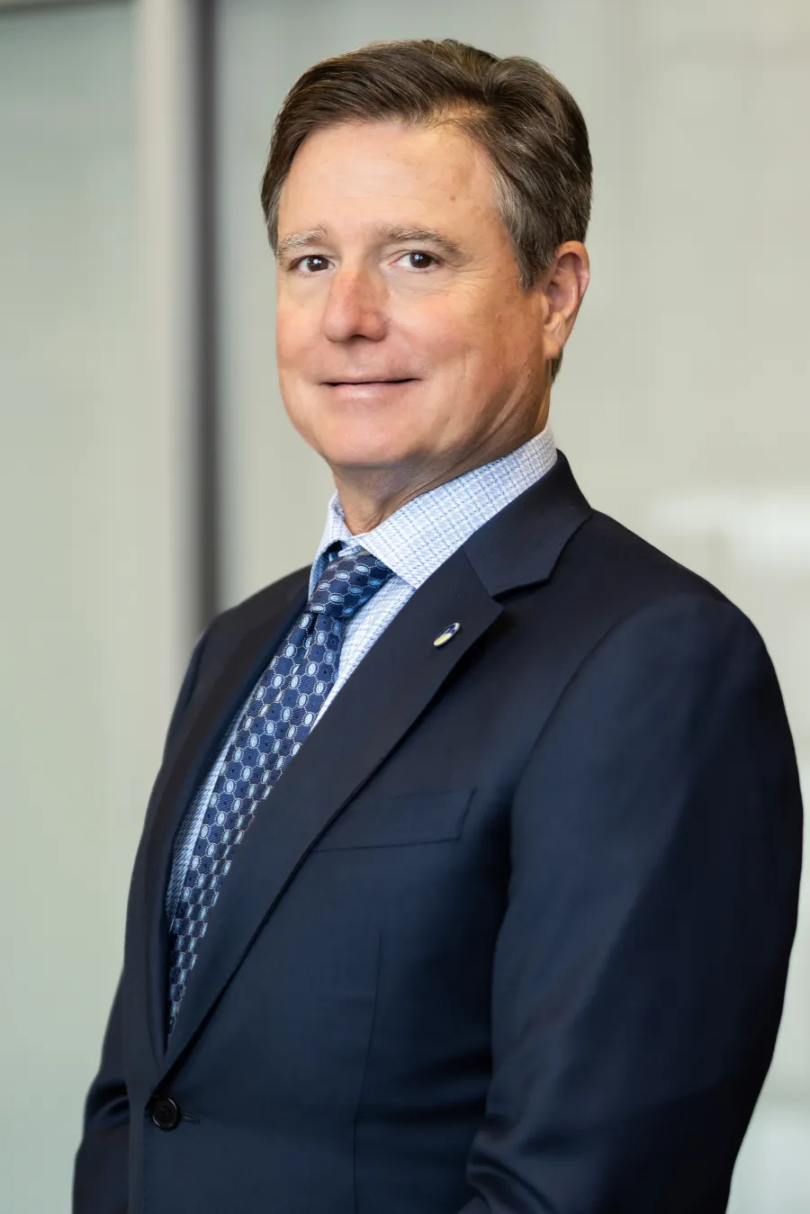 Professional middle-aged man in dark suit and patterned tie posing in office setting with neutral background