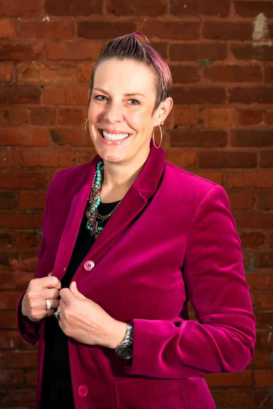 Smiling woman with short hair wearing a bright pink velvet jacket and turquoise necklace against brick wall.
