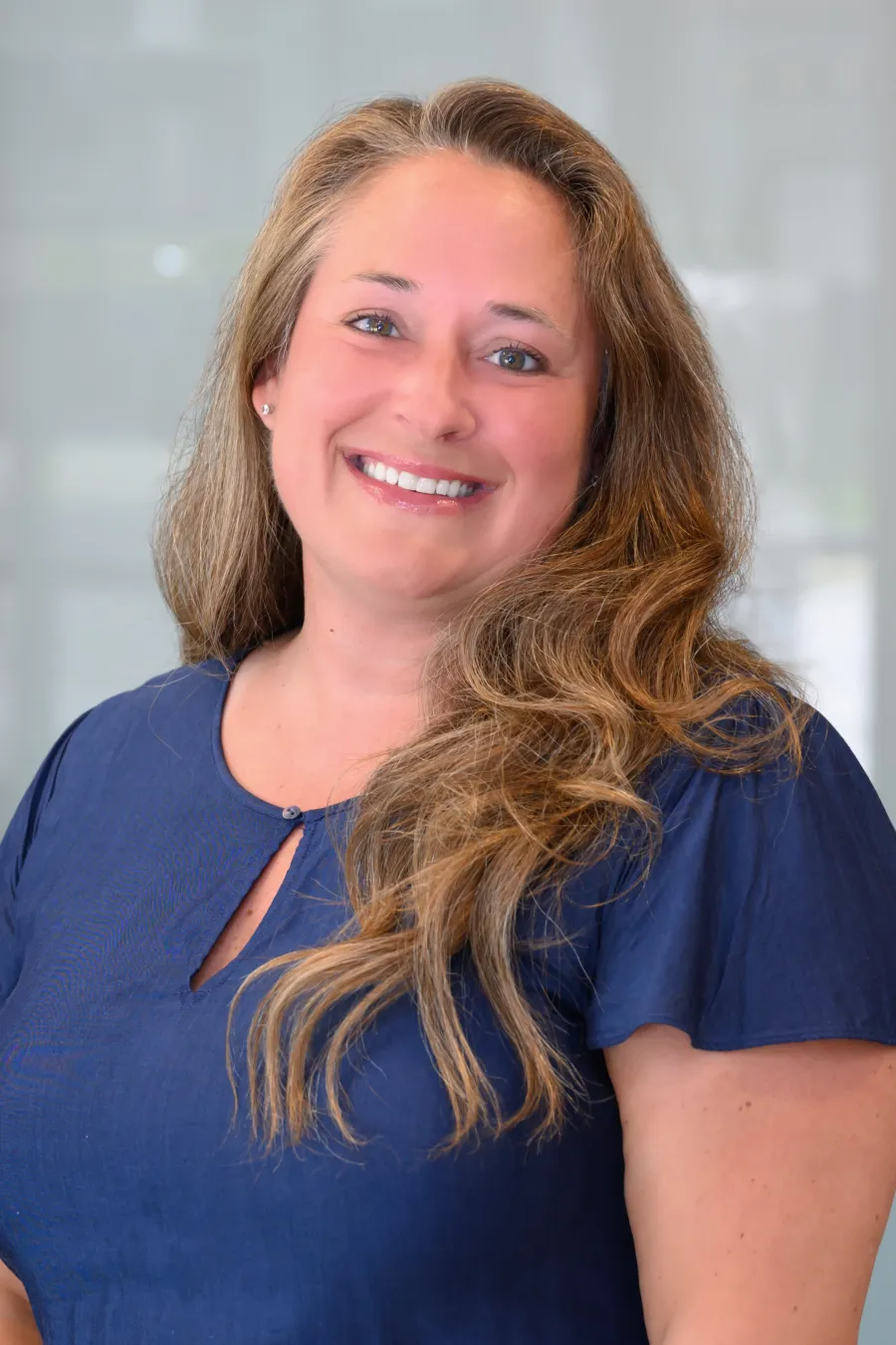 Smiling woman with long wavy hair wearing a navy blue blouse against a blurred background