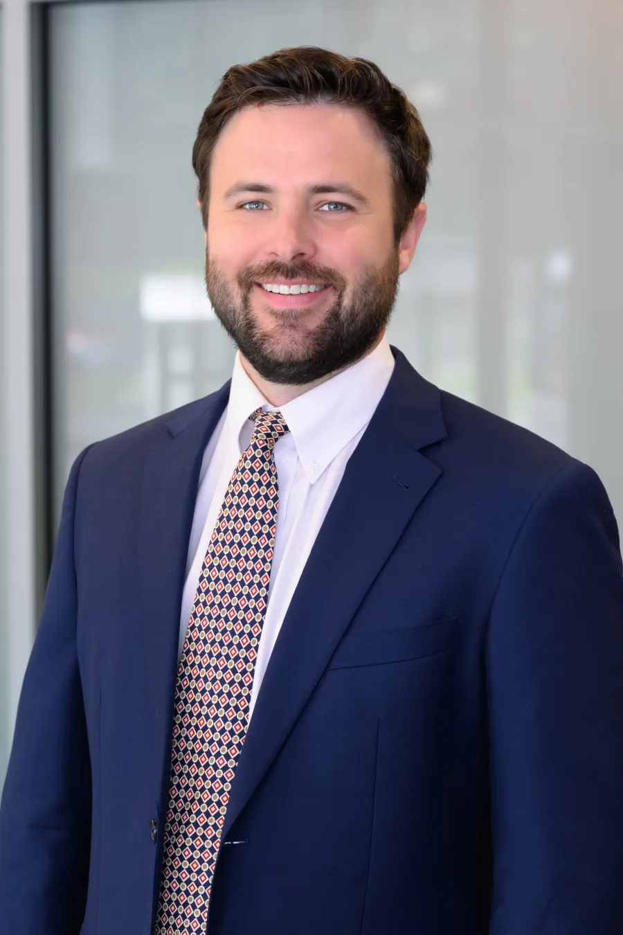 Smiling man with beard wearing a navy suit and patterned tie standing in a bright office environment.