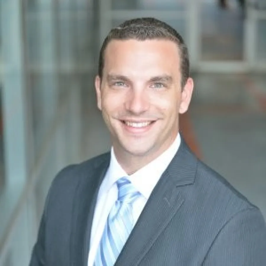 Smiling man with beard wearing a suit jacket and collared shirt with blurred greenery background