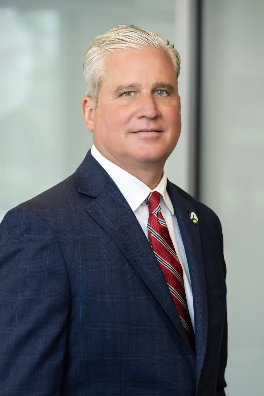 Professional portrait of a man in a navy checkered suit with a red striped tie against a blurred office background