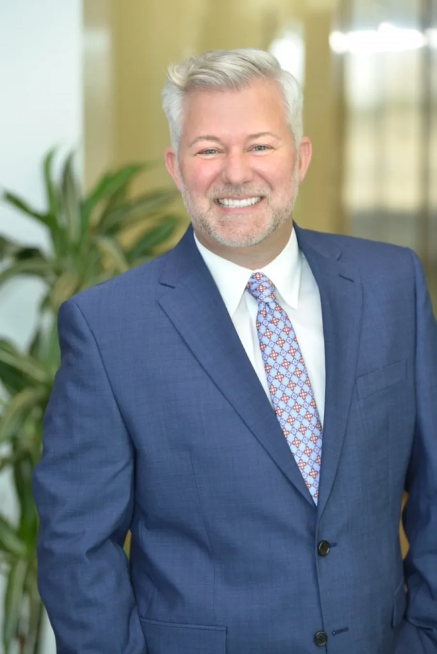 Smiling middle-aged man with gray hair in blue suit and patterned tie standing indoors with plants in background.