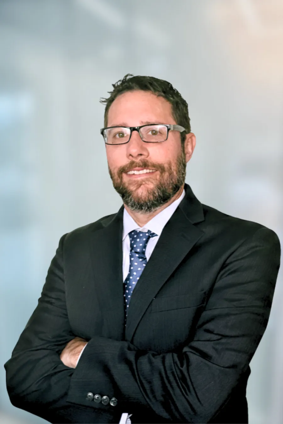 Professional man with beard and glasses wearing a dark suit and blue polka dot tie in an office.