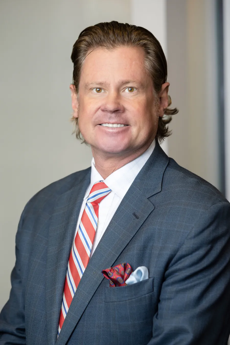 Smiling man in gray suit, white shirt, and red striped tie with pocket square in professional indoor setting.