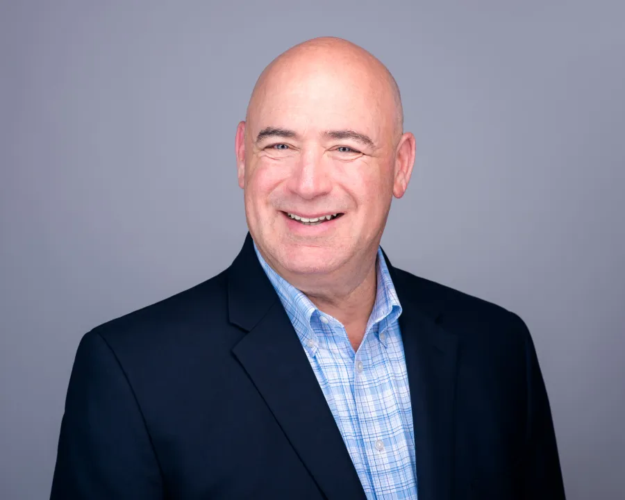 Smiling bald man in business attire wearing a checkered shirt and dark blazer against a gray background