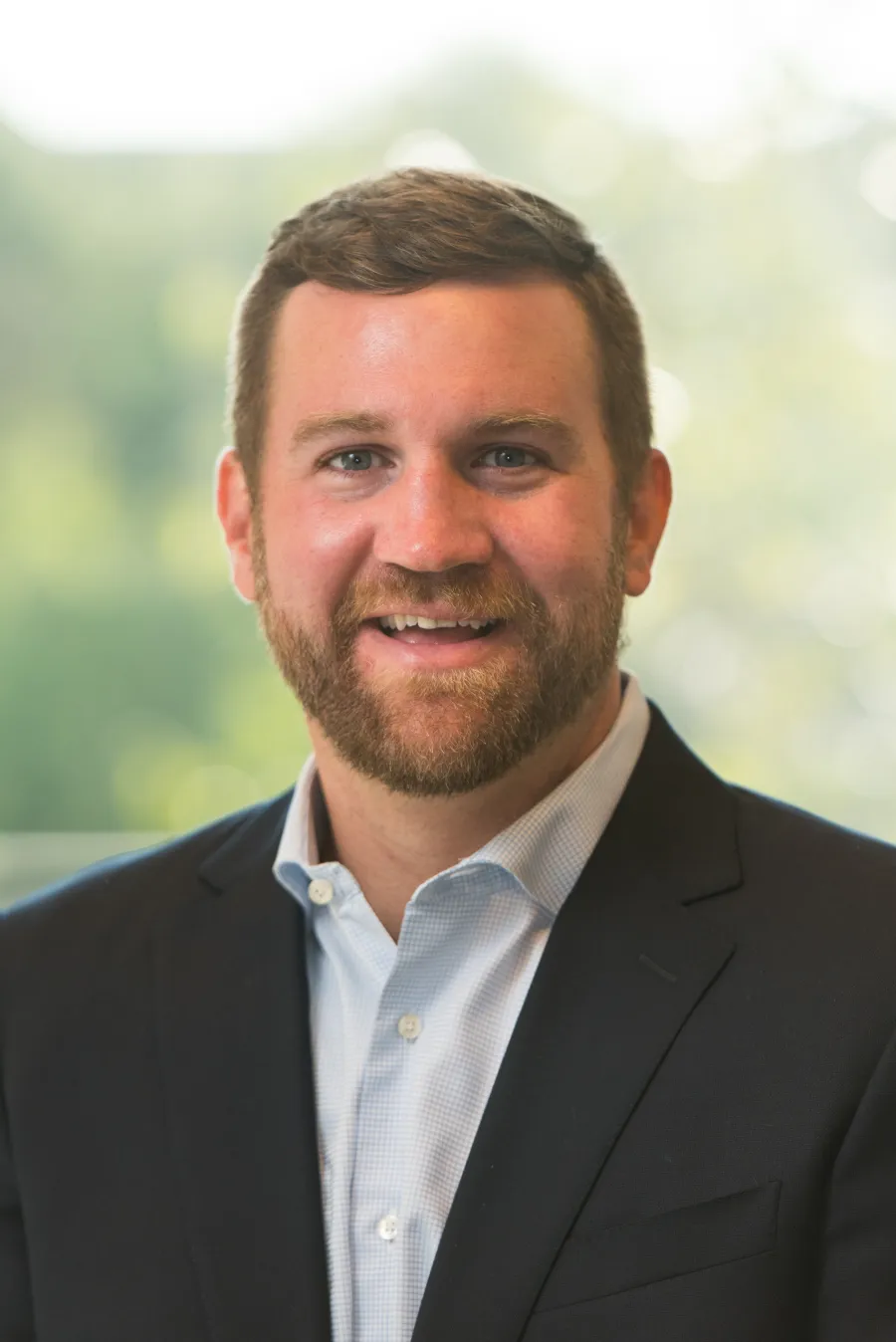Smiling man with beard wearing a suit jacket and collared shirt with blurred greenery background