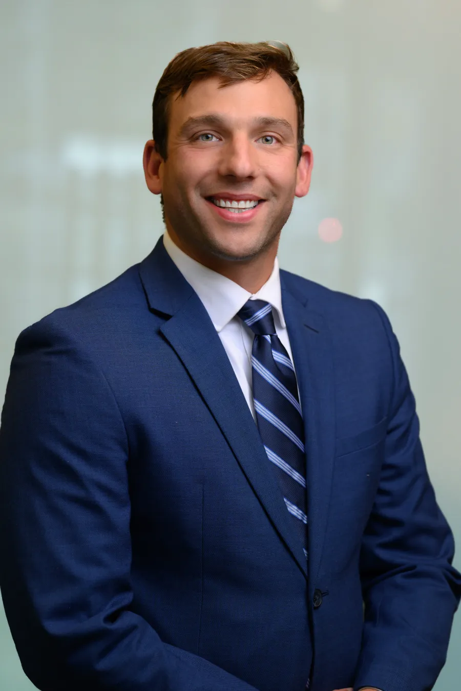 Smiling young man wearing a navy blue suit and striped tie, posing against a blurred background.