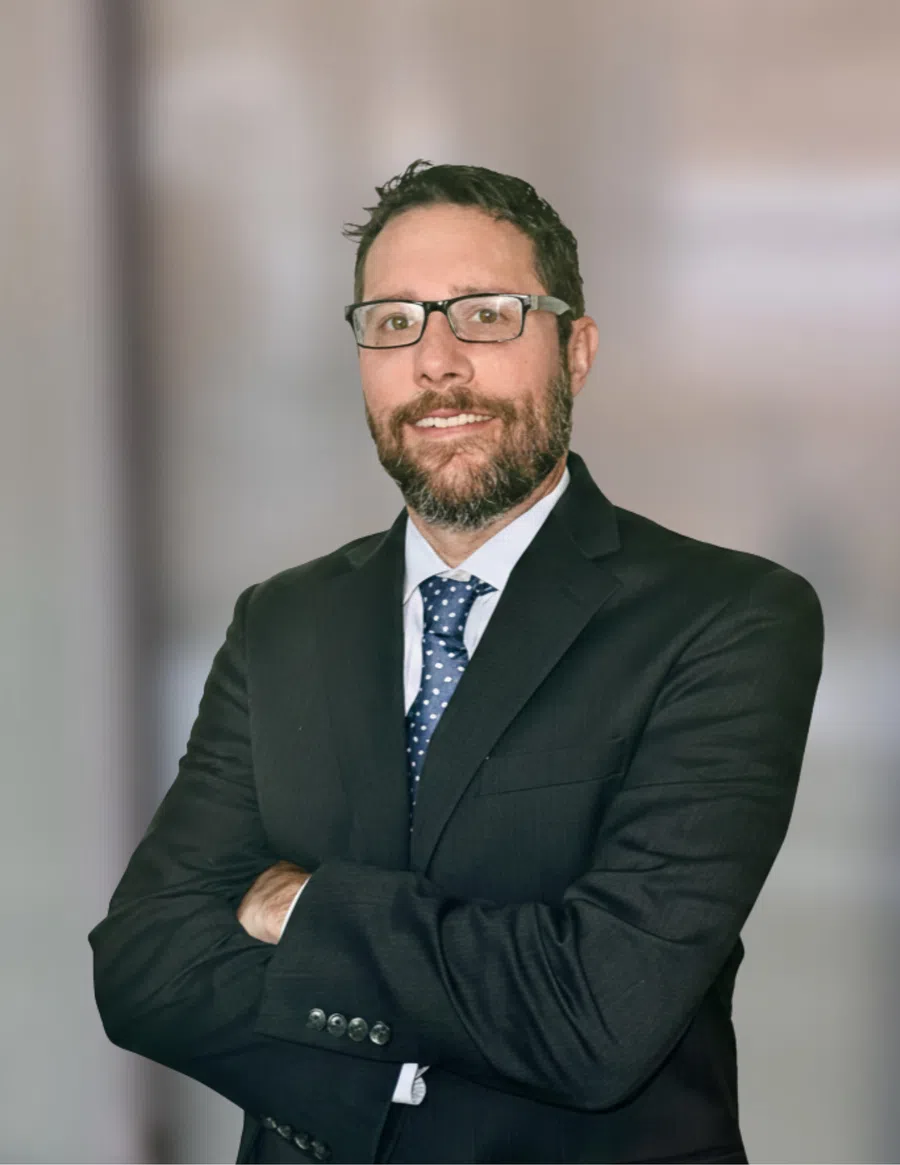 Smiling man with glasses and beard wearing a dark suit and blue polka dot tie standing with arms crossed.