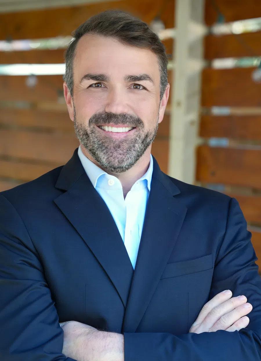 Smiling man in a navy suit with folded arms standing against a wooden wall background