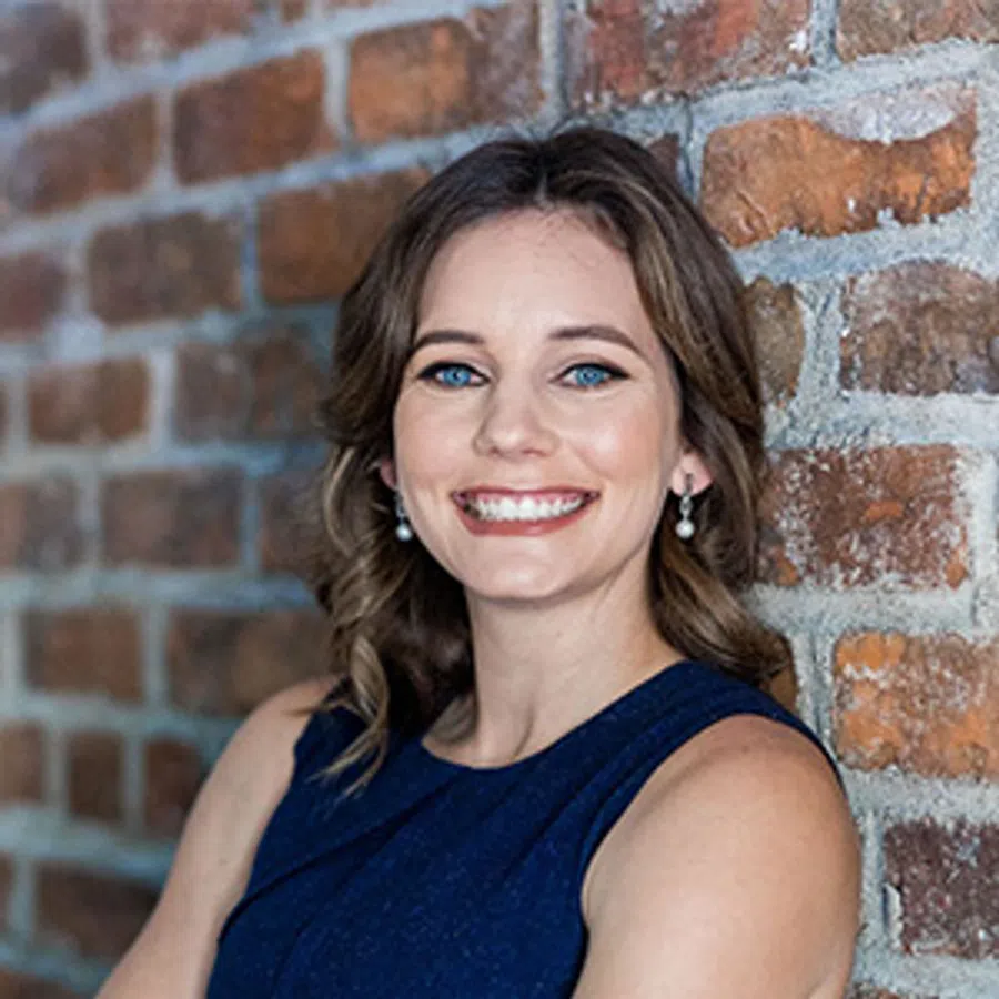 Smiling woman with blue eyes and pearl earrings wearing navy dress leaning against brick wall