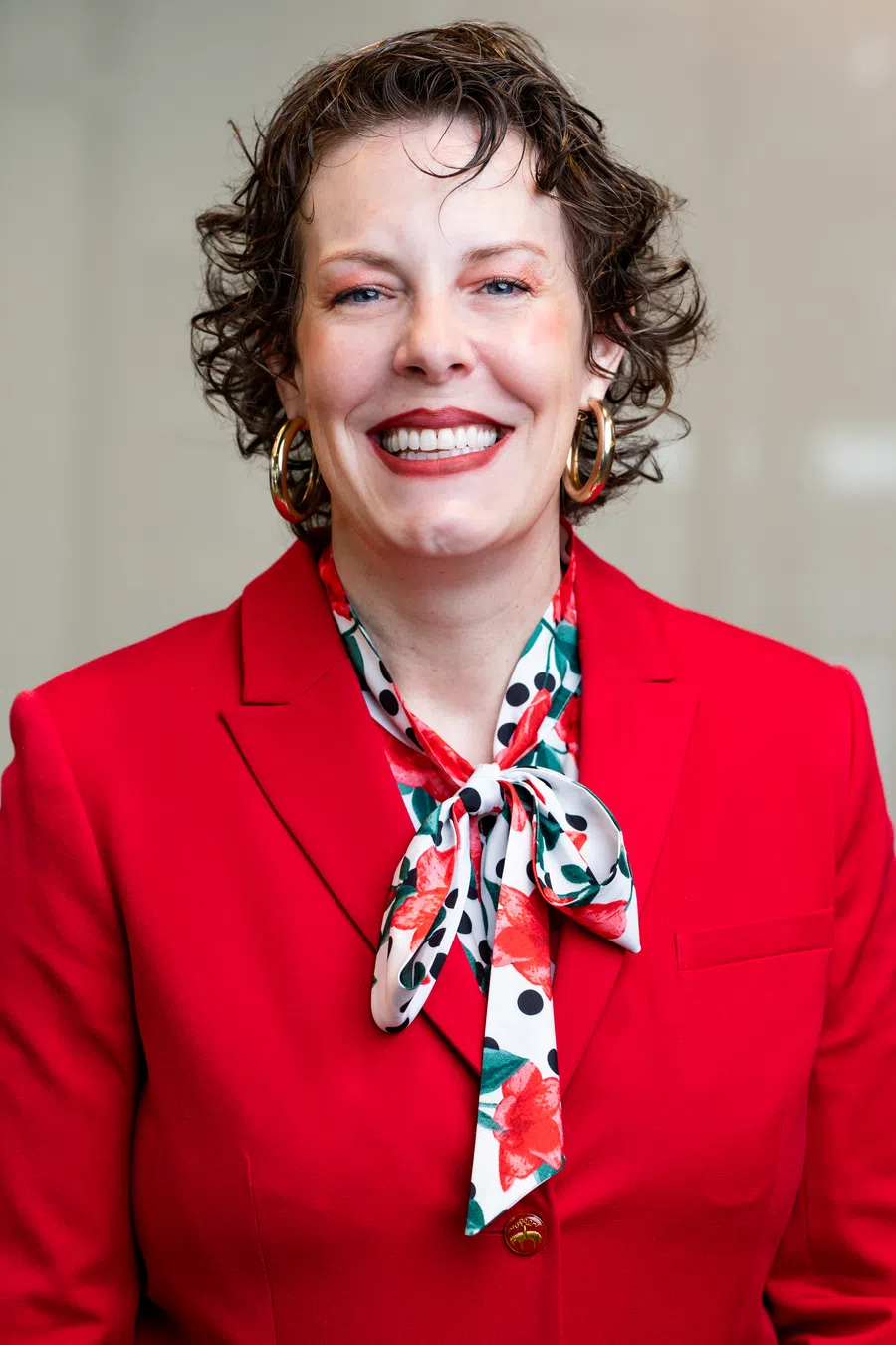 Smiling woman with curly hair wearing a red blazer, floral polka dot scarf, and gold hoop earrings.