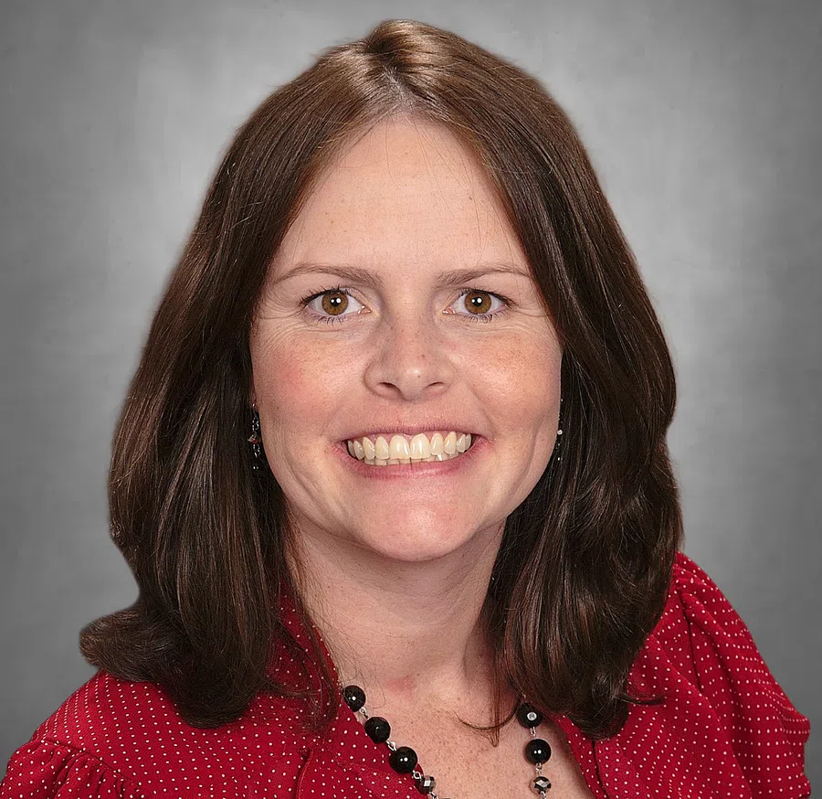 Smiling woman with medium brown hair wearing a red polka dot blouse and black beaded necklace against gray background