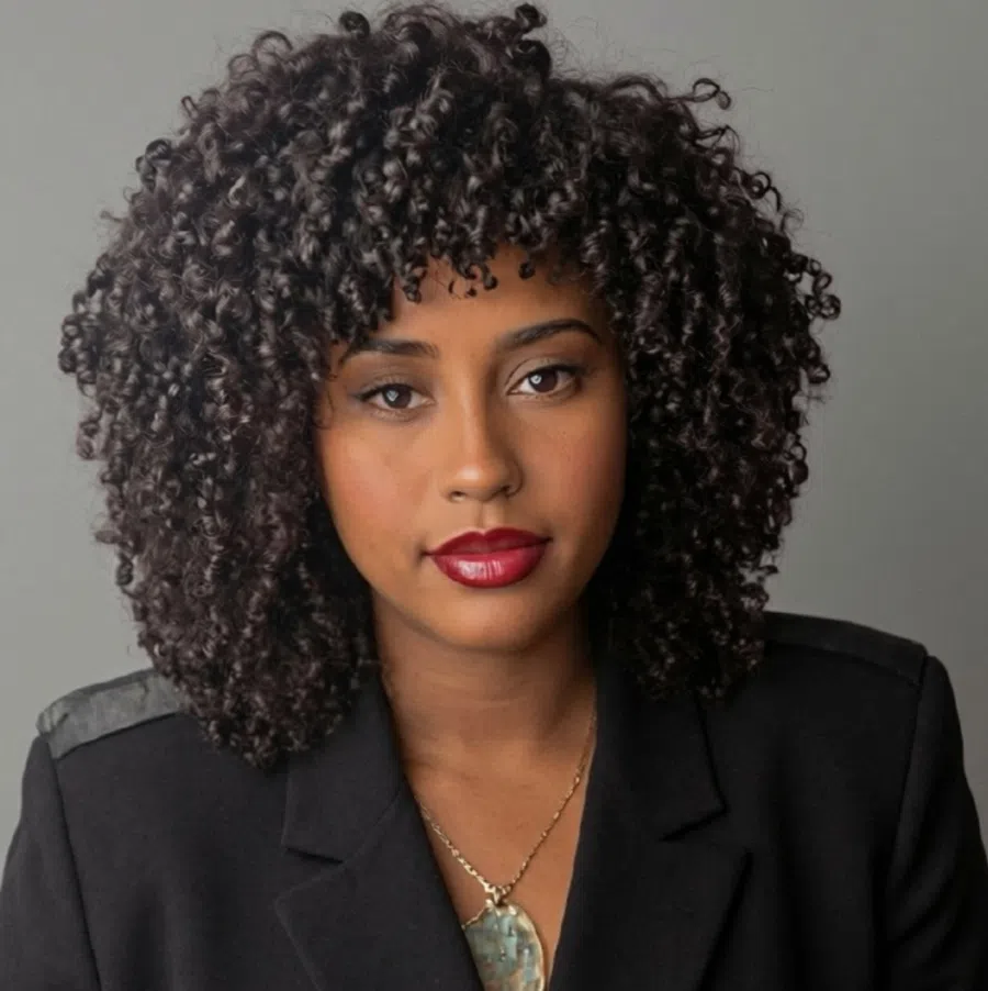 Portrait of a woman with voluminous curly hair, red lipstick, wearing a black blazer and pendant necklace.