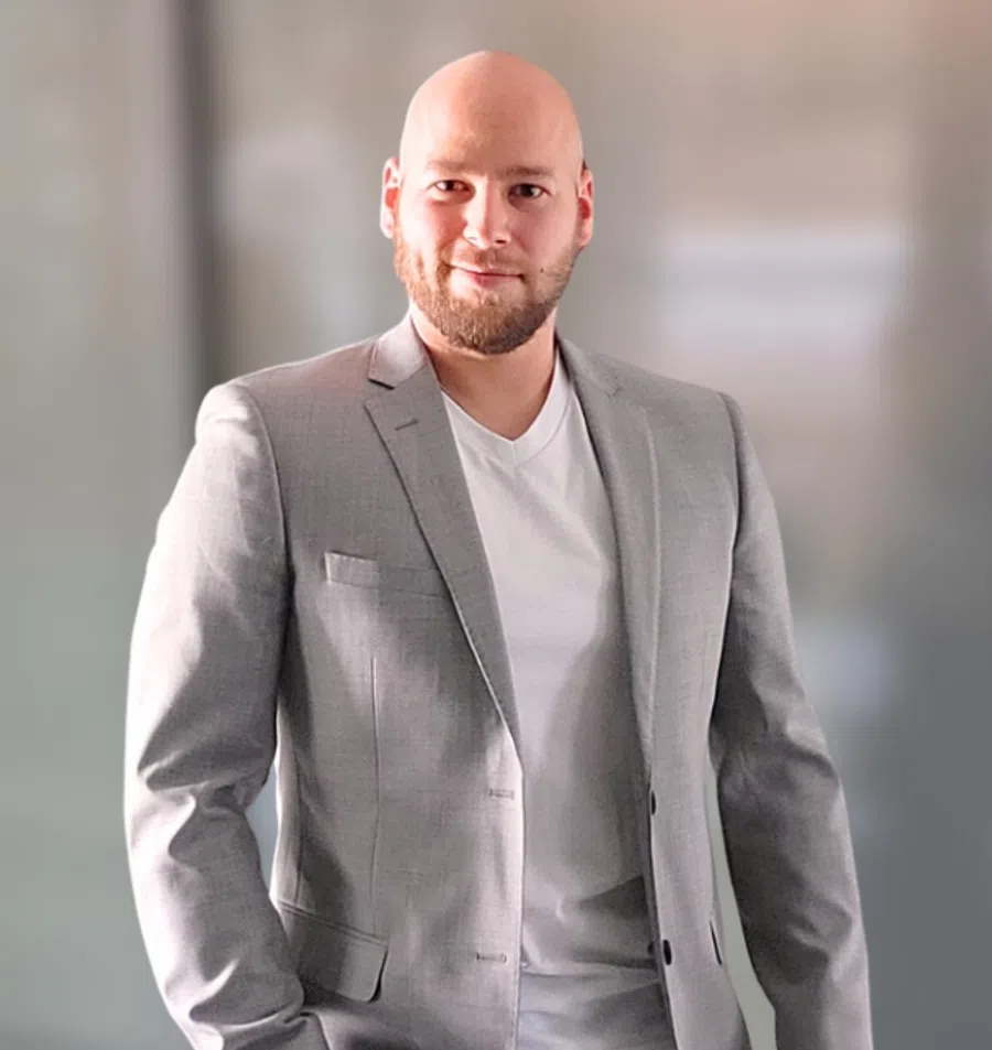 Confident bald man with beard wearing gray suit jacket and white t-shirt against blurred background