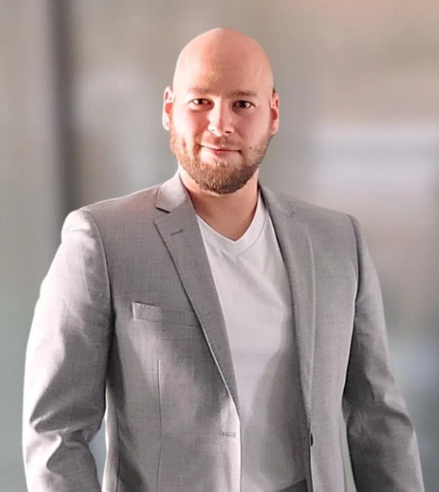 Smiling bald man with beard wearing a light gray suit jacket and white shirt against a blurred background