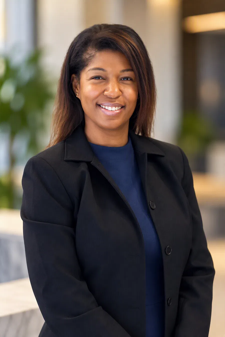 Confident professional woman in black blazer smiling in a modern office setting with blurred background.