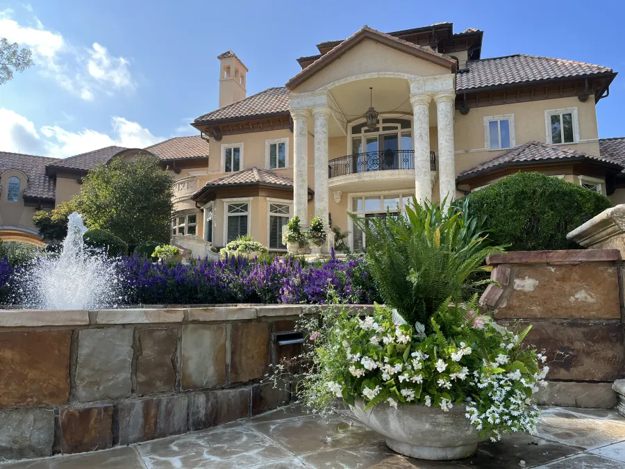 Luxurious mansion with tall columns, landscaped garden, vibrant flowers, and a stone water fountain under blue sky.