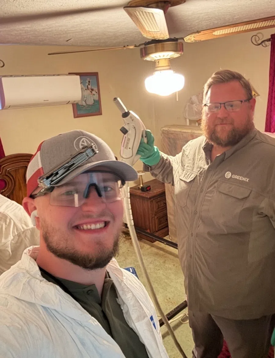Two pest control workers in protective gear inside a room, one holding a spray device and both smiling at the camera.